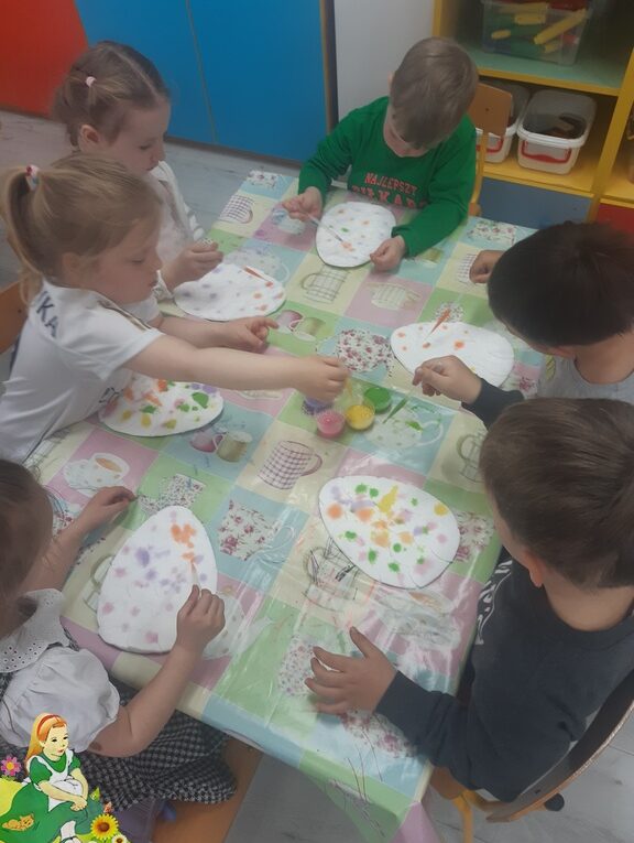 Dzieci przy stolikach malują pisanki za pomocą pipet z farbami.